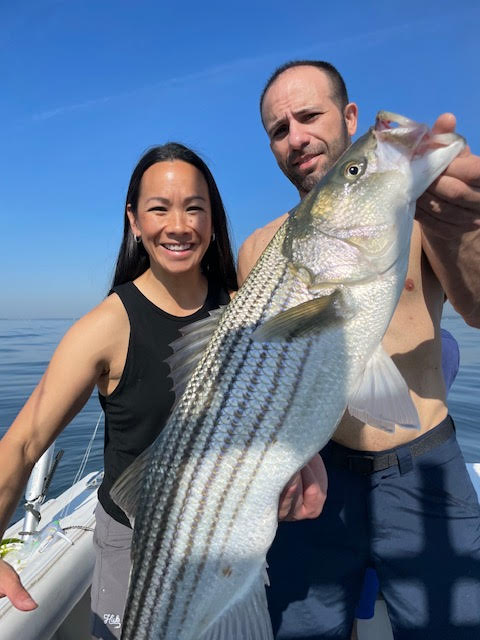 (PHOTO: Nuno Decosta with his wife Karen and a striped bass they caught this season using the Reliable Bunker Spoon lure in a glow pattern sold exclusively at Tyalure.)