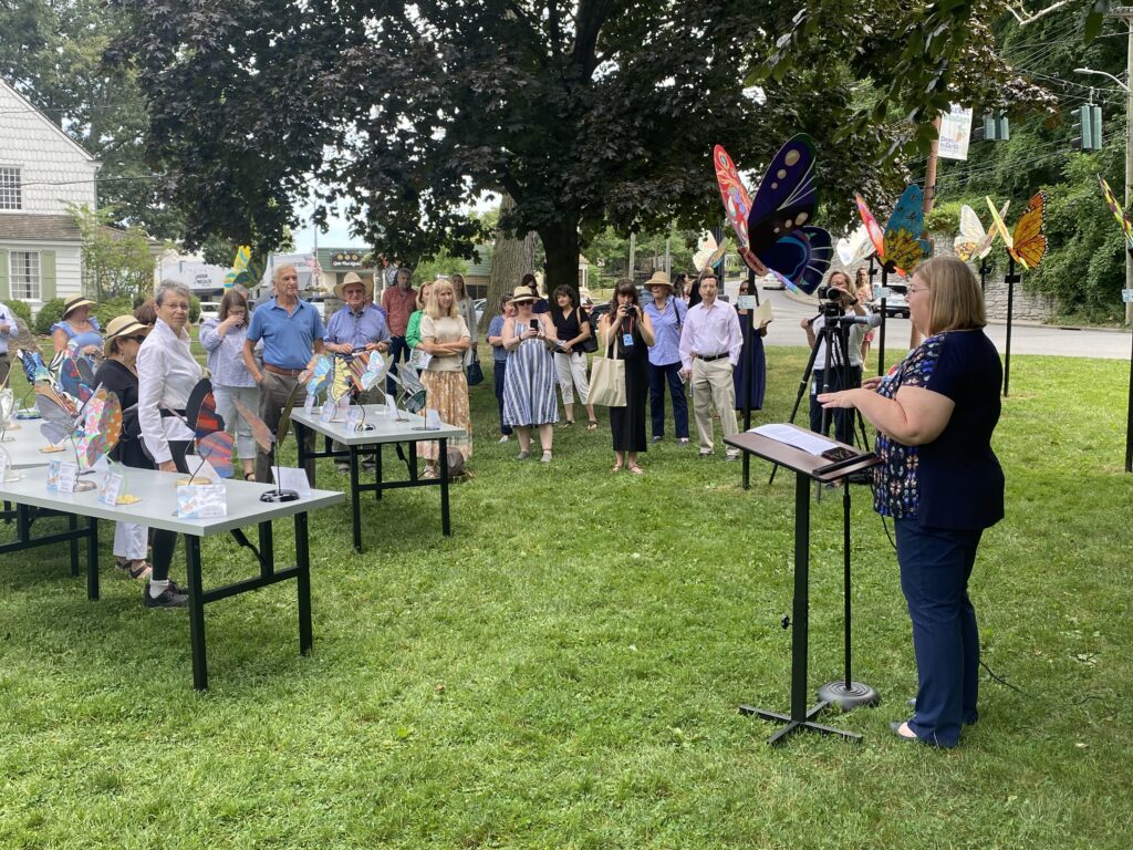 (PHOTO: Michele Thomas, president of Rye Rotary, addresses the assembled crowd at the unveiling of the Rye’sAbove butterflies on the Village Green.)