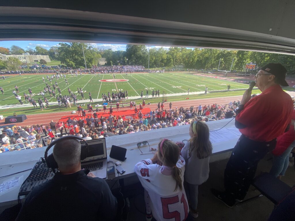 (PHOTO: From the Nugent Stadium press box, Steve "the OG" Feeney (standing, in red) calls the game, while Kevin Devaney Jr. (sitting, far left) broadcasts onto the LocalLive network and two lucky young fans enjoy a great view at the Rye Harrison football game on Saturday, October 8, 2022.)