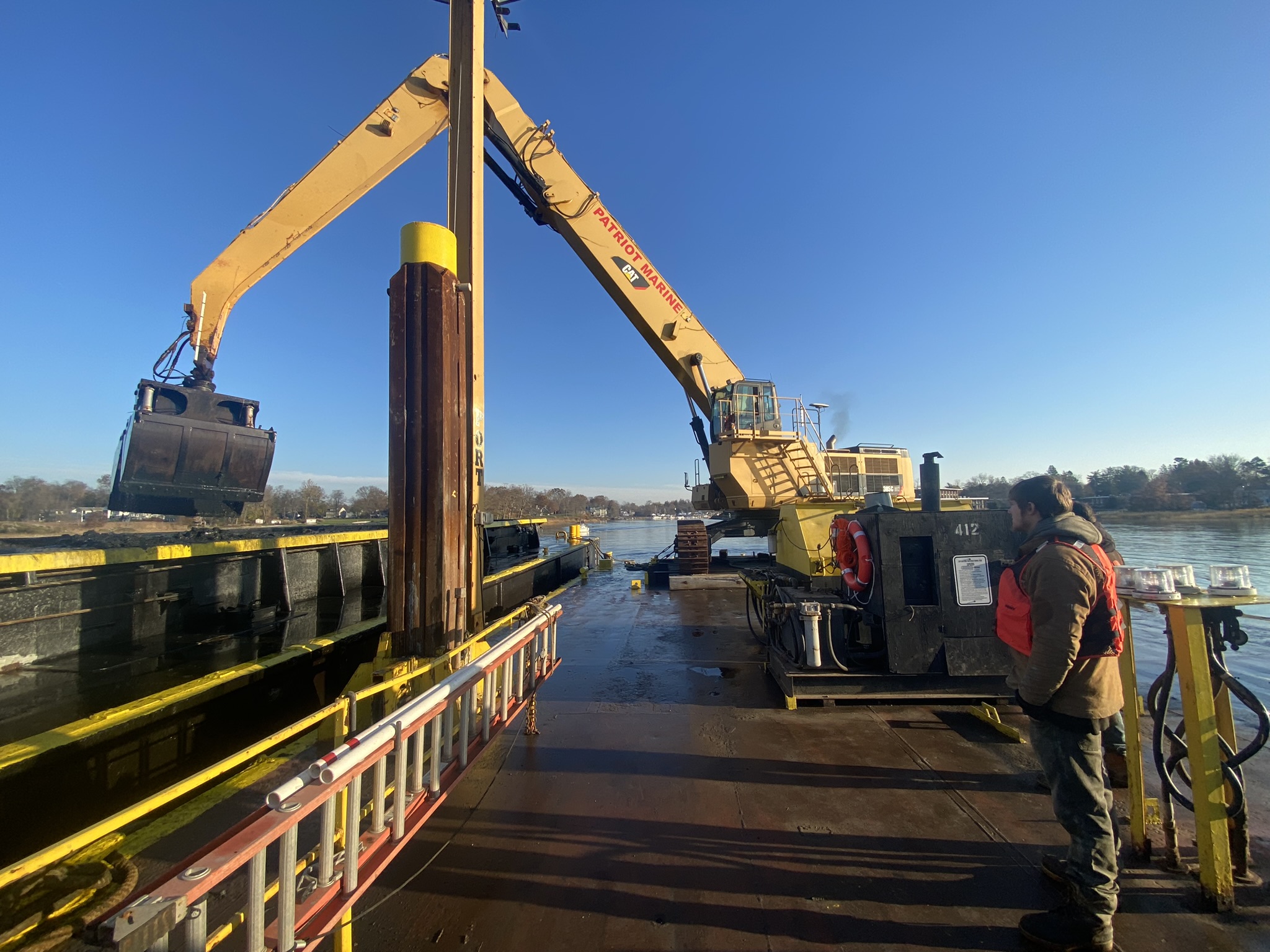 (PHOTO: Dredging operations last Wednesday in Milton Harbor. The crane operator removes each bucket of silt and transfers it to the scow on the left.)