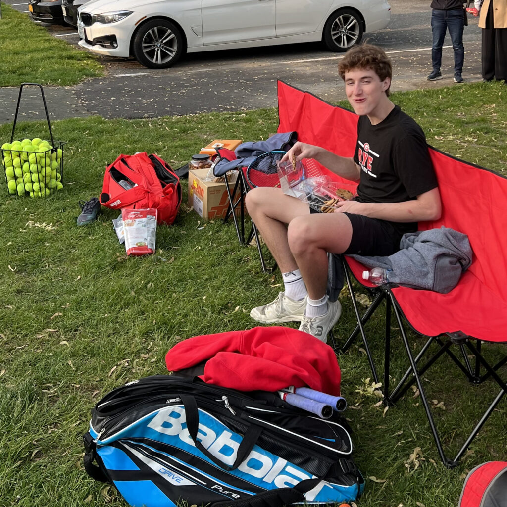 (PHOTO: Rye Boys Varsity Tennis player Henry Bagley with a postgame snack on the bench.)