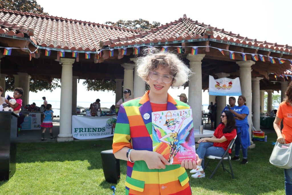 (PHOTO: Desmond Napoles posing with their book “Be Amazing, a History of Pride” after the read-aloud in Rye Town Park on Saturday, June 10, 2023. Credit: Sierra Desai.)