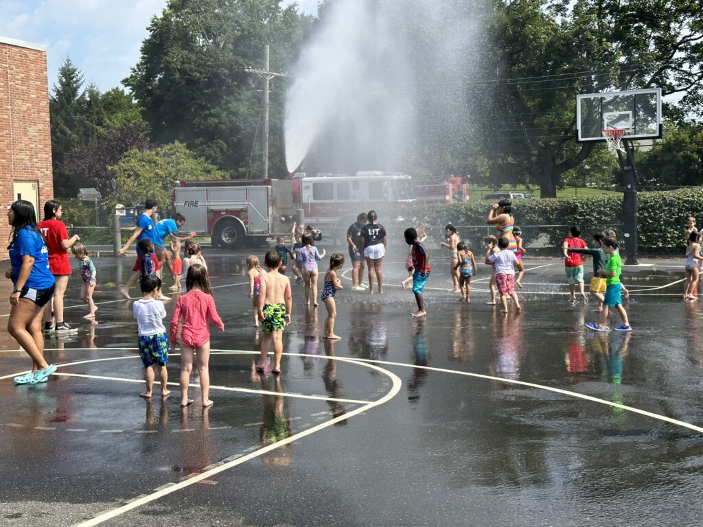 (PHOTO: On Thursday, July 20, 2023, the Rye Fire Department brought their firetruck to the Rye Y's Discovery Camp to cool down the Explorers group campers  (ages 4 & 5). The camp is located at the Osborn School.)