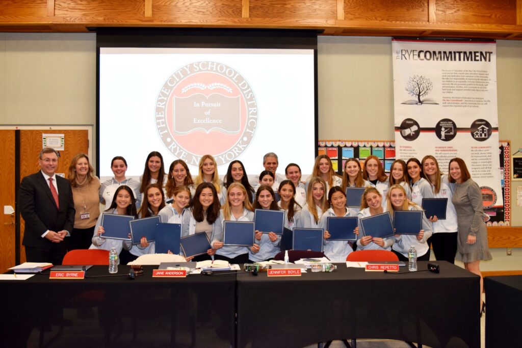 (PHOTO: The champion Rye High School girls varsity soccer team received Rye Recognition of Excellence Awards from Superintendent Dr. Eric Byrne at the December 5, 2023 Board of Education meeting. On hand to congratulate the girls were County Executive George Latimer, Lisa Urban, Chief of Staff to New York State Representative Steve Otis, and Rye Mayor Josh Cohn. The photo shows (most of) the team with Coach Rich Savage, Dr. Byrne, Athletic Director Susan Reid Dullea, and Principal Suzanne Kelly Short.)