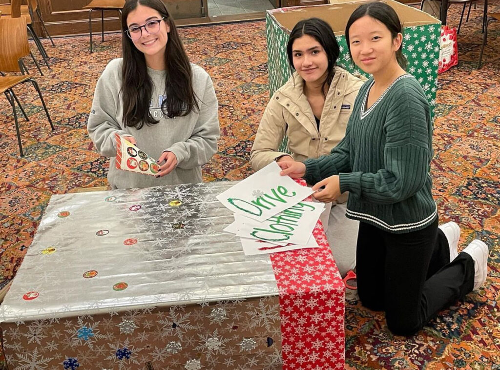 (PHOTO: Rye High School senior Paulina Tepan, winner of the 2023 Rye Youth Human Rights Award, in the middle, along with Alexis Friedman (left) and Grace Wang (right), setting up a box for the Knight’s Boutique clothing drive at Rye High School.)