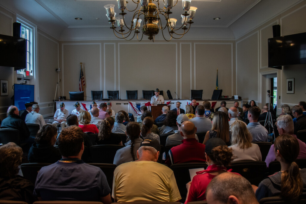 (PHOTO: Commander Frederick de Barros speaks in front of a crowded City Hall chambers on Memorial Day 2024. Credit: Tilman Oberbannscheidt.)