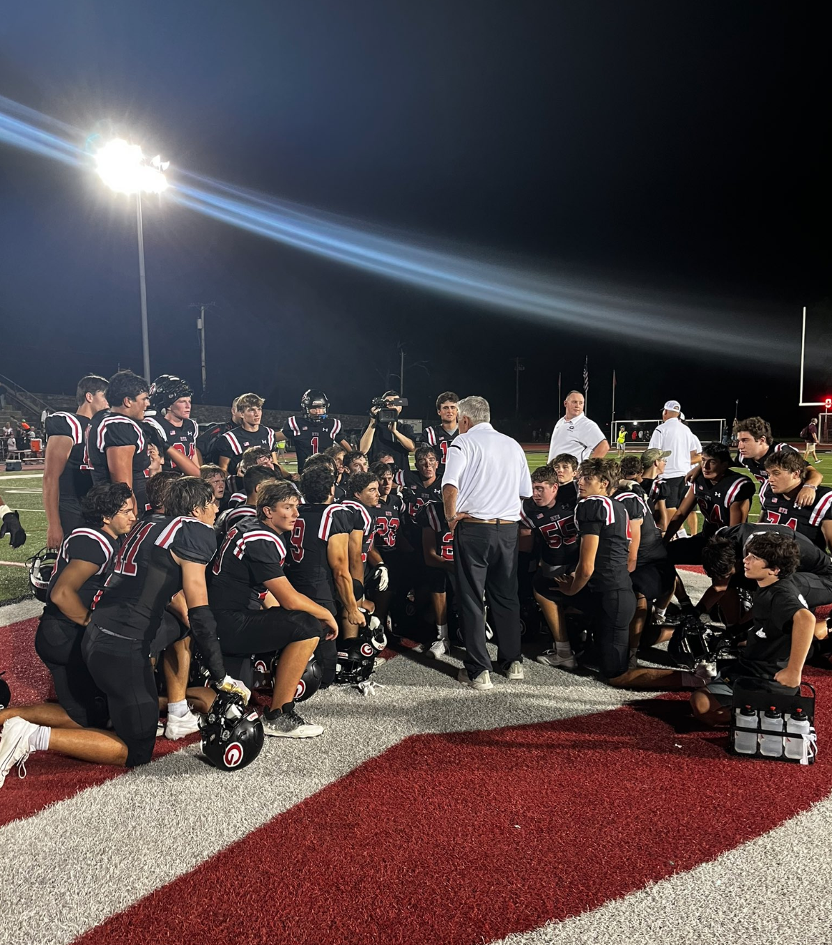 (PHOTO: Rye huddles around head coach Dino Garr after its 31-21 Week 0 win. Credit: @RyeAthletics on X)