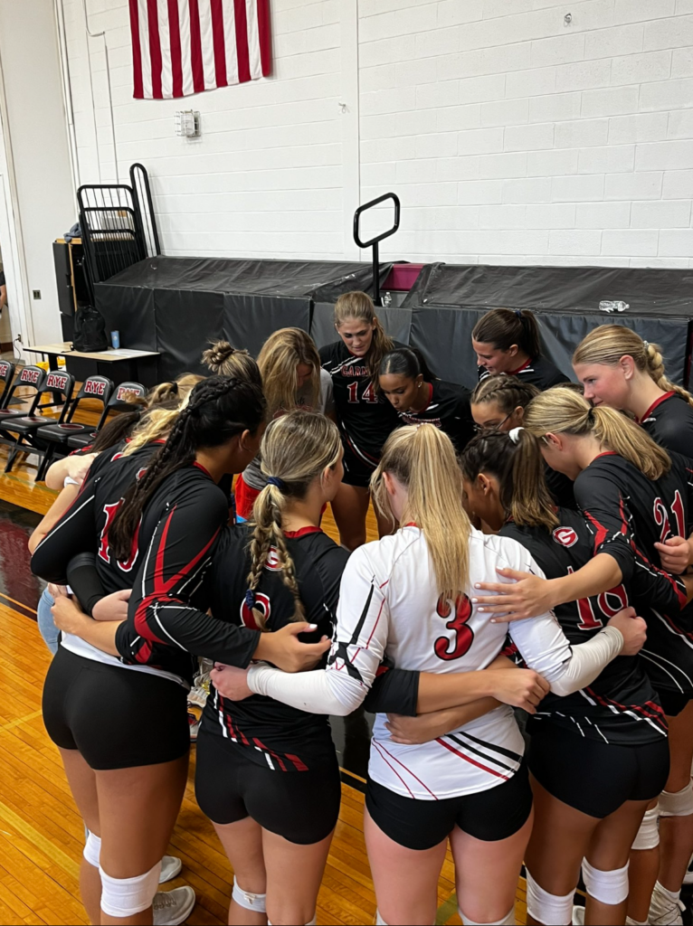 (PHOTO: Rye Varsity Volleyball huddles around head coach Geri Jones after taking a 2-0 lead in sets against Nanuet on September 6, 2024.)