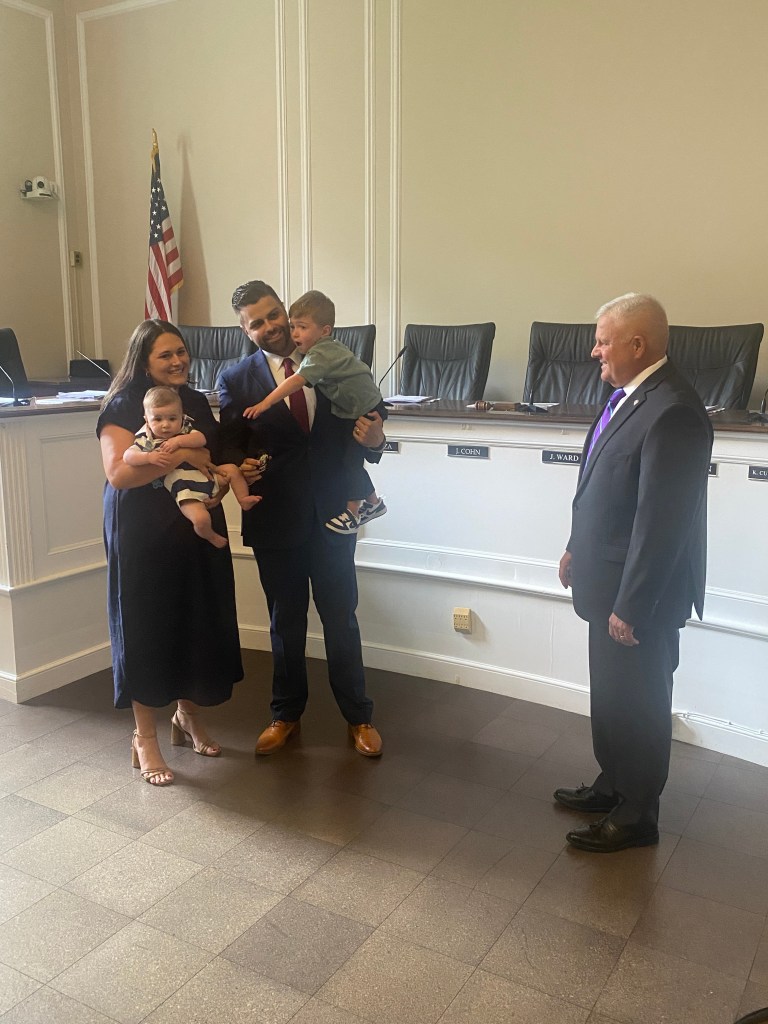 (PHOTO: Detective Robert Jones with his wife Christina, baby Rocco (6 months) and son Logan (3 years). Public Safety Commissioner Mike Kopy looks on at Jones' crowd control skills at Jones' promotion at Rye City Hall on September 17, 2025.)