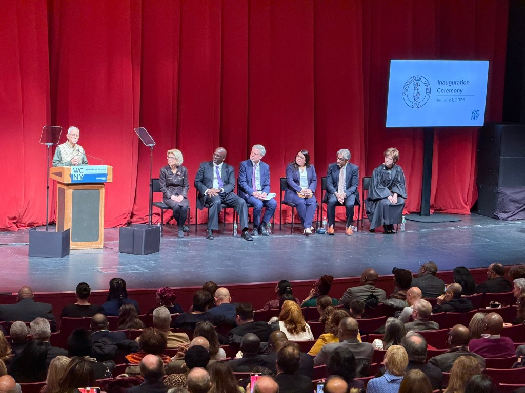 (PHOTO: Arun Bhagirath from the Hindu Temple of Tri-State in White Plains delivering one of the closing benedictions at the Westchester County inauguration event on January 1, 2026 at the Performing Art Center at Purchase College. Seated left to right: State Senator Shelley, County Executive Ken Jenkins, County Clerk Thomas Roach, County Legislators Jennifer Puja and Anant Nambiar and the Honorable Anne E. Minihan.)