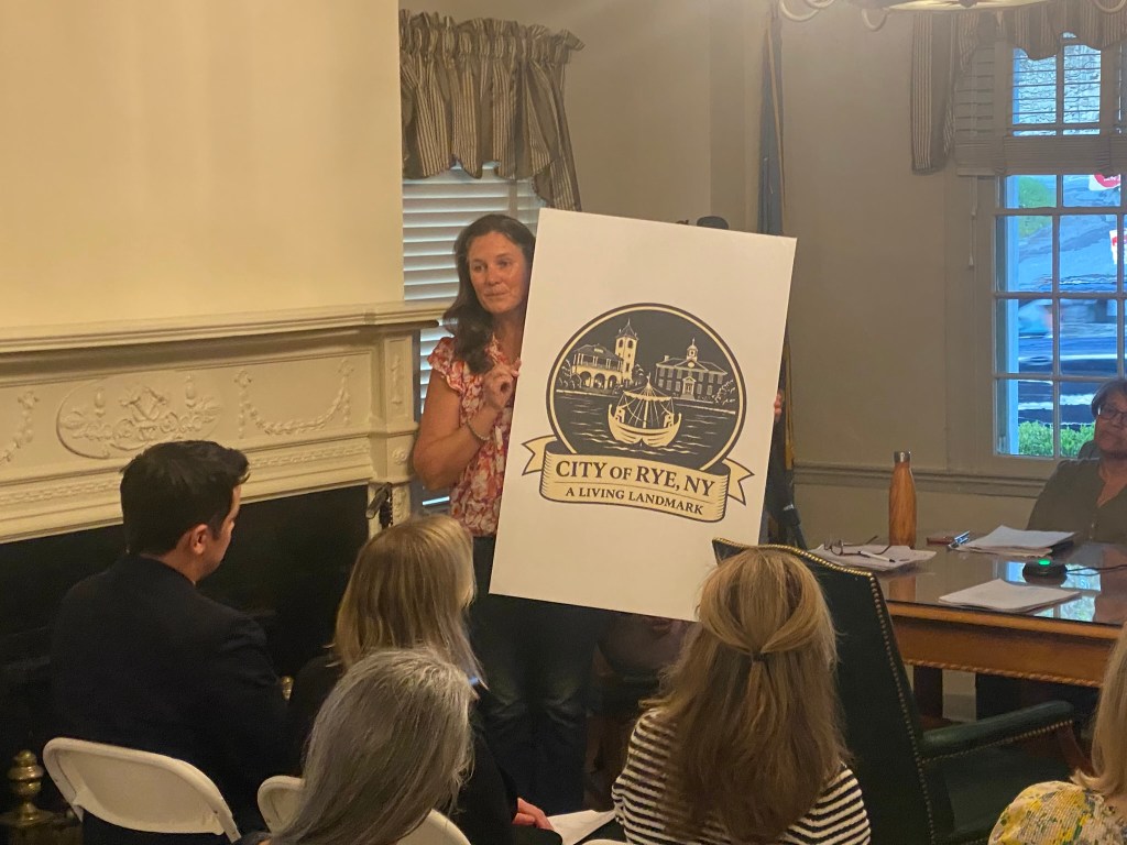 (PHOTO: New Landmarks Advisory Committee Chair Alison Cupp Relyea shows a new logo showing Rye as a "living landmark" to be used around the America 250 celebrations to celebrate Rye and its people. Relyea was addressing  the annual Square House meeting of the Rye City Council on April 15, 2026.)