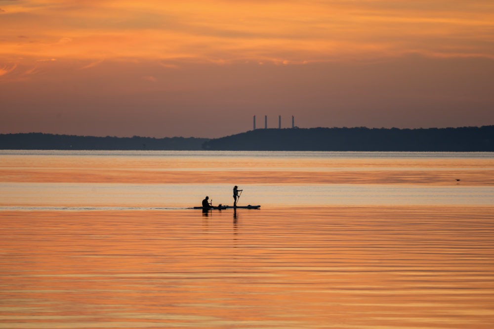 (PHOTO: Setting out at Sunrise. Enveloped in a wash of orange dawn light, a kayaker and paddleboarder glide quietly across the calm water. By Stacey Massey, author of First Light, Rye.)