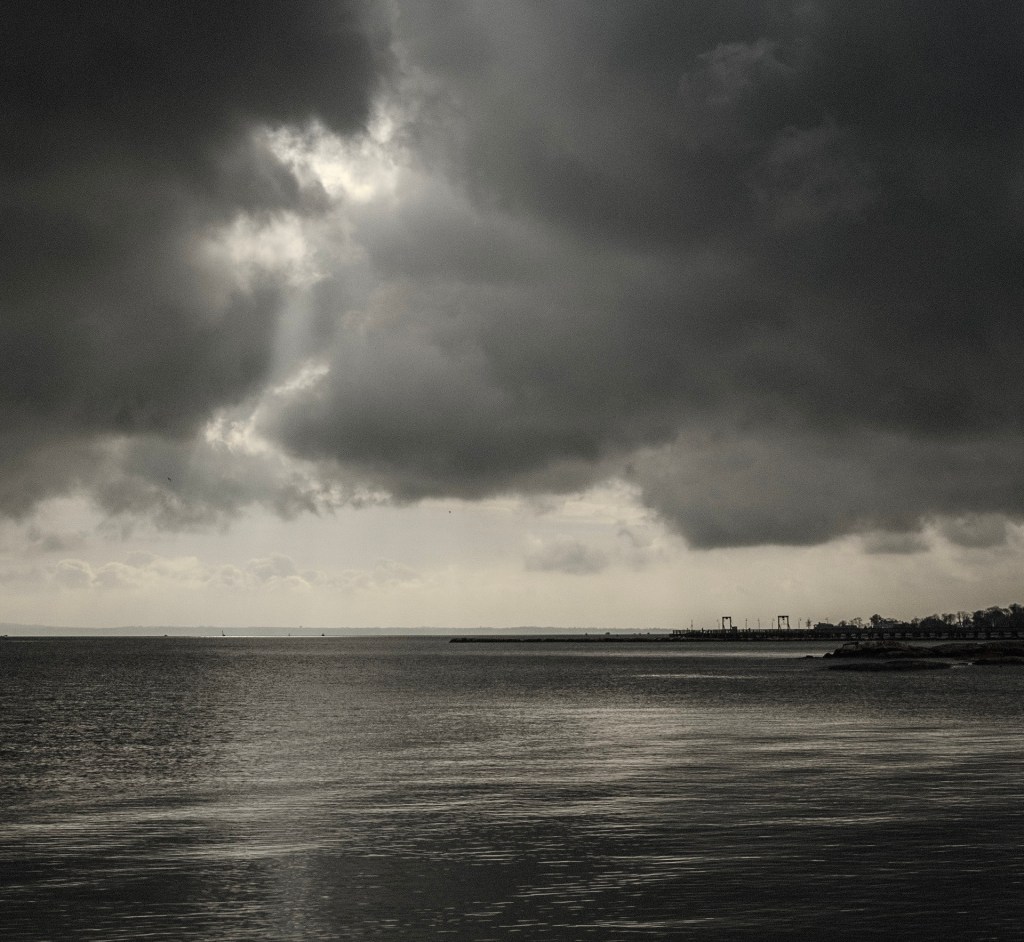 (PHOTO: Breakthrough. Sunlight splitting dense clouds to spill out over the Long Island Sound. By Markus Peters.)